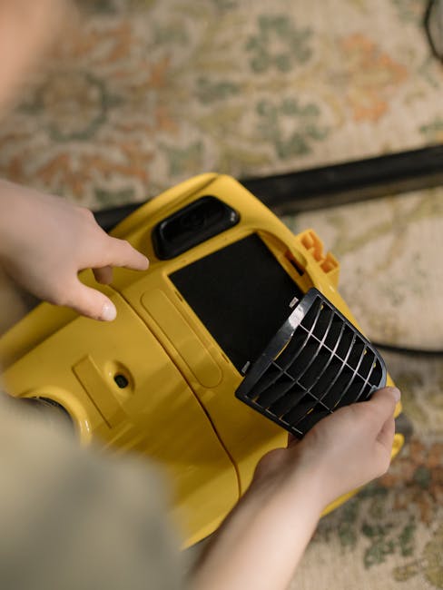 A woman kneeling on a patterned rug in a living room with warm lighting, placing a compact vacuum cleaner with a black and yellow design onto the carpet. The vacuum's transparent dust container reveal