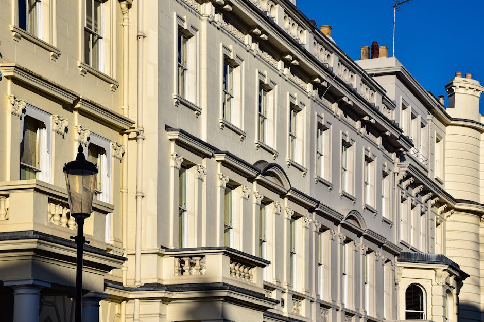 A close-up view of an elegant, cream-colored Victorian-style building with ornate architectural details, multiple sash windows with white frames, decorative mouldings, and small balcony railings, illu