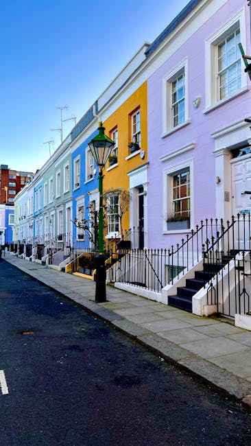 A row of colorful terrace houses on a residential street, featuring pastel-colored facades in shades of purple, yellow, and light blue. Each house has a small front garden with black wrought iron railings and steps leading up to white-framed windows and doors. A vintage-style street lamp is situated on the sidewalk, which is paved with concrete slabs. The scene is well-lit with natural daylight, highlighting the clean and well-maintained appearance of the buildings and pavement, reflecting the importance of surface cleaning and upkeep in domestic and commercial properties, as promoted by Carpet Cleaning SW7 for end of tenancy cleaning quotes in SW7.