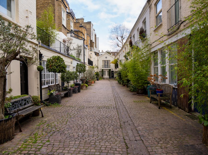 A narrow cobblestone alleyway in a residential area during daytime, flanked by white buildings with large windows and black wrought iron balconies. Potted plants and small trees line the sides, with wooden benches and greenery creating a pleasant, tidy outdoor space. The alley is well-lit with natural light, and the scene conveys a clean, maintained environment consistent with high-end urban housing. The setting suggests a peaceful, well-kept courtyard or garden area, typical of residential streets in Kensington. Carpet Cleaning SW7 specializes in surface cleaning and deep cleaning services, ensuring hygiene and cleanliness for similar residential spaces.
